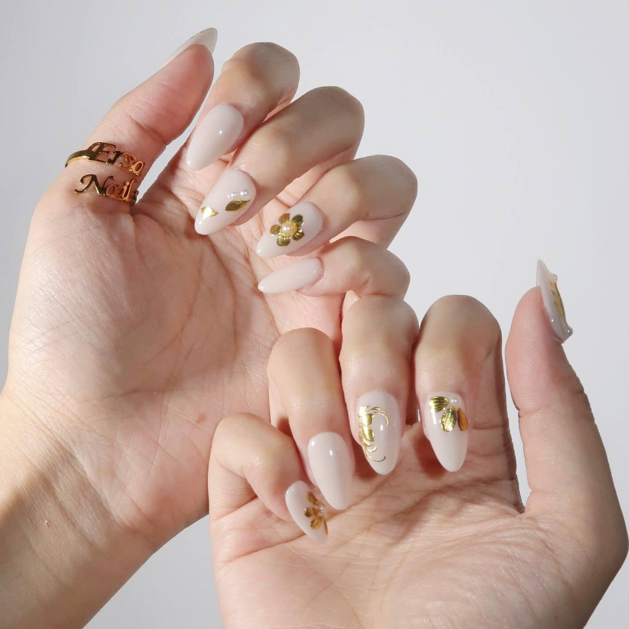 Hand with beige nails and gold accents, wearing a gold ring, on a light background