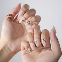 Thumbnail for Hand with beige nails and gold accents, wearing a gold ring, on a light background
